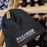 Schwarze Strickmütze mit dem Schriftzug 'MARX Städter' und Hashtag '#Chemnitzerbier' auf einem Holzregal mit Flaschen.